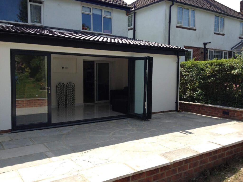 A modern house extension with large glass folding doors opens onto a stone patio, leading to a sunny backyard, bordered by a low brick wall and hedges.