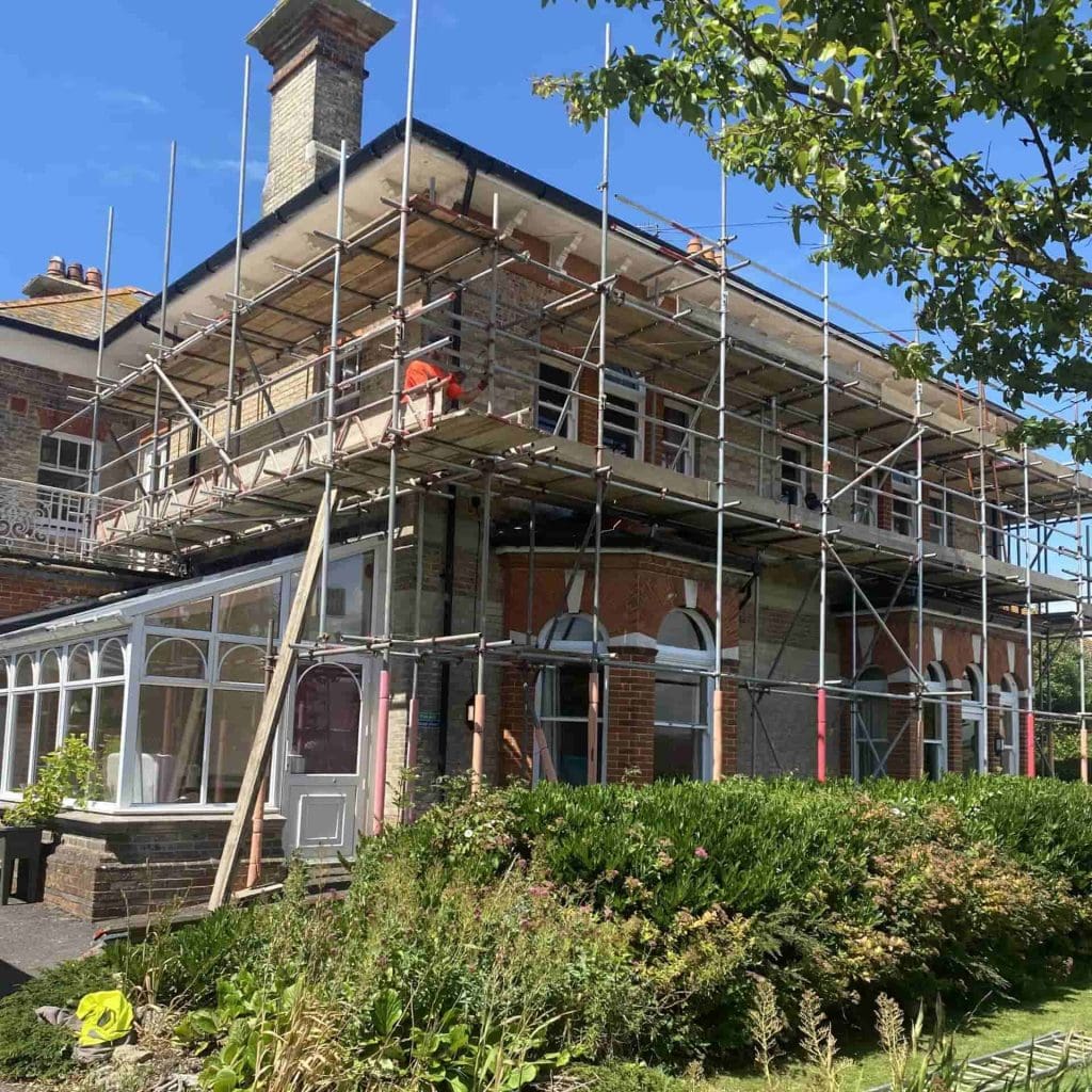 A large brick house surrounded by scaffolding, with a person working on the upper level. There is a glass conservatory attached to the side and lush greenery in the foreground under a clear blue sky.